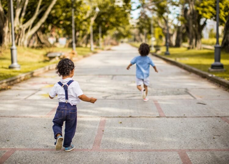 Two young children running joyfully on a paved pathway in a scenic park setting, enjoying a sunny day.