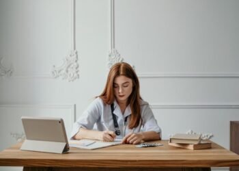 A woman doctor writing notes at a desk with a tablet and books, capturing a professional office setting.