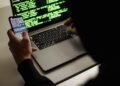 From behind crop male programmer in black hood browsing netbook and mobile phone while working in office