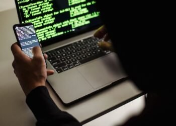 From behind crop male programmer in black hood browsing netbook and mobile phone while working in office