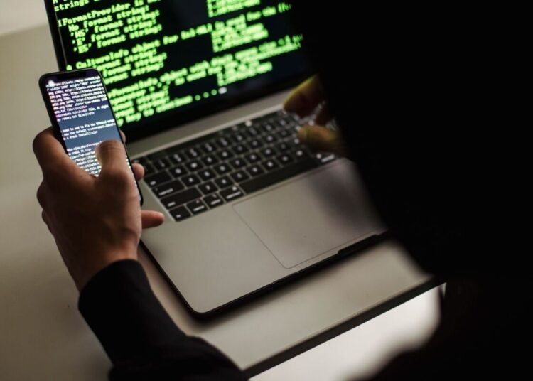 From behind crop male programmer in black hood browsing netbook and mobile phone while working in office