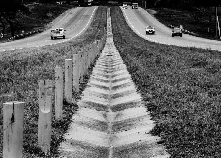Monochrome view of a highway with cars and a wooden fence in the foreground.