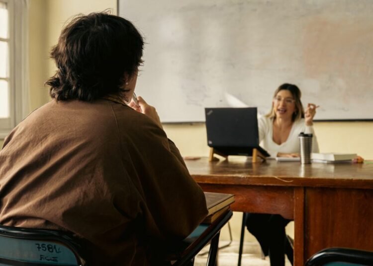 Student and teacher engage in discussion in a classroom setting with a laptop and whiteboard visible.