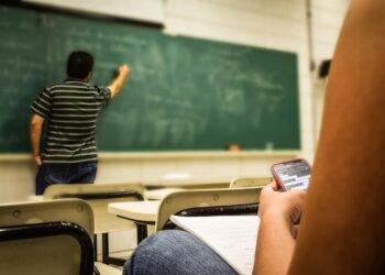 Student texting in a classroom while teacher is writing on the blackboard.