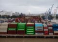 Aerial view of colorful shipping containers in a bustling port with cranes and city skyline.