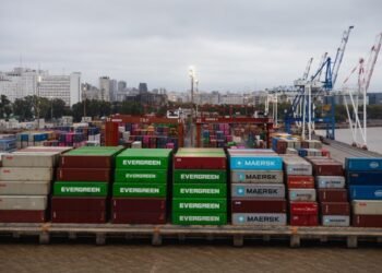 Aerial view of colorful shipping containers in a bustling port with cranes and city skyline.