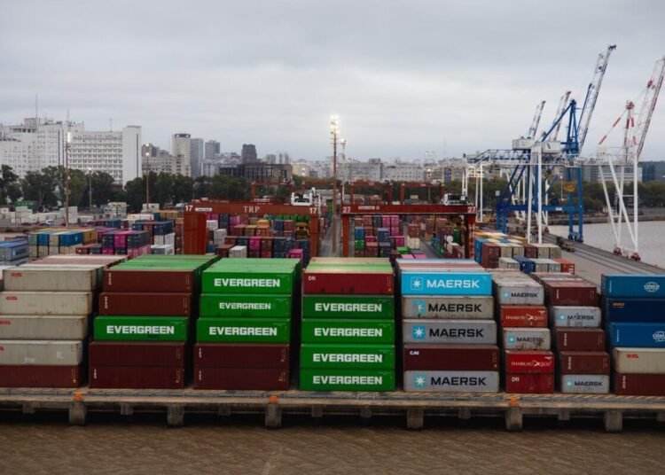 Aerial view of colorful shipping containers in a bustling port with cranes and city skyline.