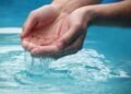 Close-up of hands scooping clear blue water from a reflective pool outdoors.