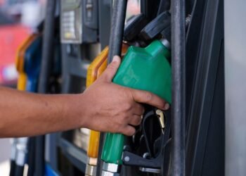 Close-up of a hand holding a green fuel nozzle at a gas pump station outdoors.